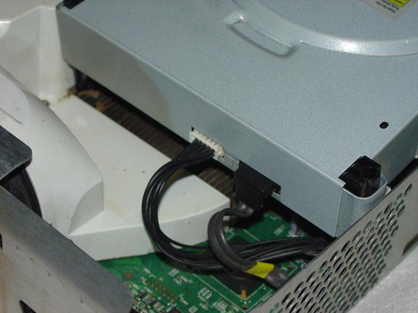 DVDドライブの前面で、最初に表示されているテープの端をはがし、DVDドライブを途中まで持ち上げて、背面の接続にアクセスできるようにします。 DVDドライブの背面にある電源とデータの接続を外し、まっすぐ持ち上げます。' alt=