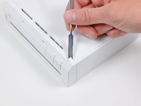 Izmantojiet metāla izsmidzinātāju, lai noņemtu baltos plastmasas skrūvju vākus, kas piestiprināti pie apakšējā korpusa netālu no Wii priekšpuses.' alt=