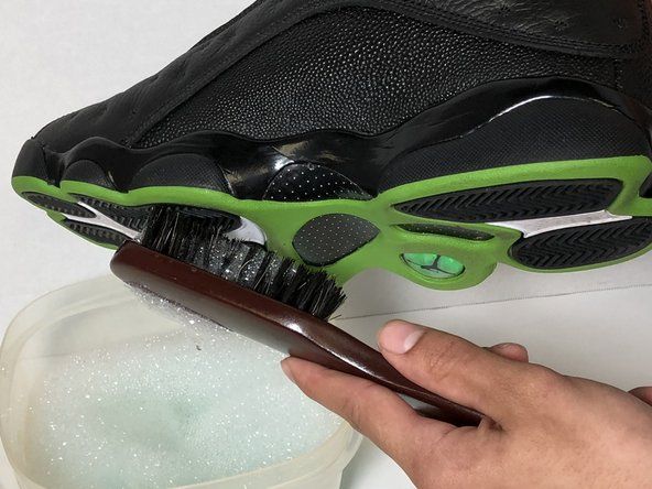 Bruk en myk børste med såpe- og vannblandingen for å skrubbe smuss fra bunnen av skoen.' alt=