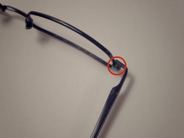 Localize o parafuso na dobradiça do canto dos óculos entre a lente e os braços e desparafuse-o usando a chave de fenda de 1,5 mm girando no sentido anti-horário.' alt=