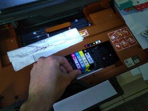 Pasiekus vagoną, spausdinimo galvutė atsidurs spausdintuvo centre. Kai jis bus centre, išimkite rašalo kasetes. Norėdami tai padaryti, paspauskite kiekvienos kasetės priekinį skirtuką. Išėmę kasetes, suvyniokite jas į popierinį rankšluostį ir padėkite į šoną.' alt=